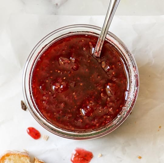 A glass jar overflowing with vibrant red homemade strawberry freezer jam, with a spoon resting inside, all set on a sheet of white parchment paper with a few drips of jam.