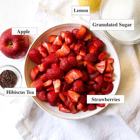 A top-down view of ingredients for strawberry jam: a white bowl filled with chopped fresh strawberries, a whole red apple, a small bowl of hibiscus, a lemon, and a glass bowl of granulated sugar, all arranged on a white linen cloth.