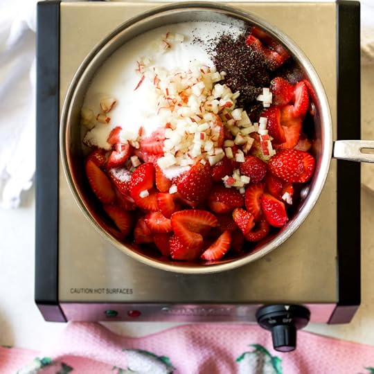 A stainless steel pot on a hot plate, filled with chopped fresh strawberries, white granulated sugar, finely diced apple, and a sprinkle of dark seasoning, ready to be cooked into jam.