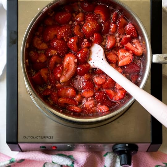 A pot of chopped strawberries, diced apple, and sugar simmering on a hot plate, with a pink spatula resting in the bubbling, macerating fruit mixture, illustrating a key step in making easy strawberry freezer jam without pectin.
