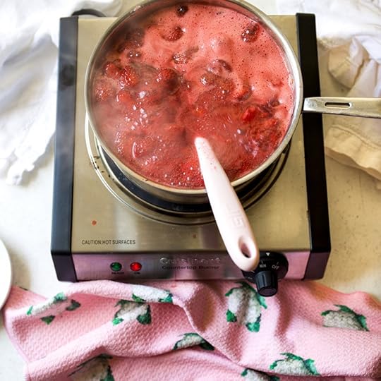 A pot of bright red strawberries, sugar, and apple rapidly boiling and bubbling on a countertop burner, with a pink spatula inside, showing the cooking stage of homemade, no-pectin strawberry jam.