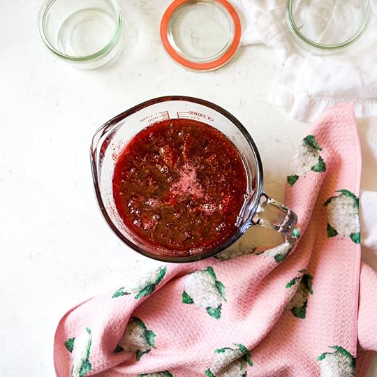A top-down view shows a glass measuring cup filled with freshly cooked, ruby-red strawberry jam, ready to be poured, with empty glass jars and a canning lid nearby, highlighting the final steps of making homemade freezer jam.