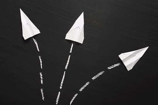 Image: three white paper airplanes lie atop a blackboard, aimed in different directions with white chalk marks behind them indicating their paths.