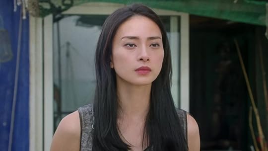 A solemn woman with long black hair stands outside a seaside structure, staring forward