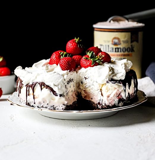 Slice removed from a homemade Neapolitan ice cream cake revealing layers of chocolate cookie crust, strawberry and vanilla ice cream, topped with chocolate ganache, whipped cream, and fresh strawberries. A container of ice cream is blurred in the background.