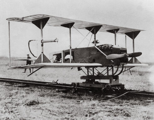 A black-and-white photo shows a large, boxy biplane sitting on tracks.