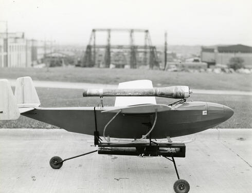 A black-and-white photo shows a small airplane sitting on a runway, with a long metal tube mounted on its back.