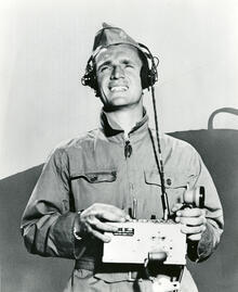 A black-and-white photo shows a man in an army uniform wearing a set of headphones and turning the dial on a large remote-control box that he is holding in front of him.