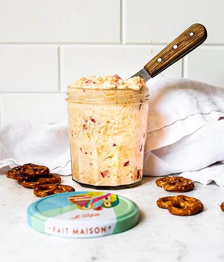 A close-up shot of creamy homemade pimento cheese in a clear mason jar with a serving knife inserted, and scattered pretzel twists on a white background.