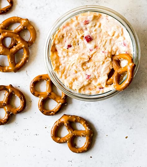 Creamy homemade pimento cheese dip in a round glass container, with several pretzel twists scattered around on a light-colored background.