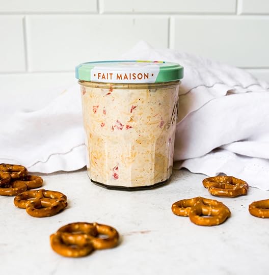 A clear glass jar filled with creamy, red-flecked pimento cheese, standing on a white surface with pretzel twists and a white napkin in the background.