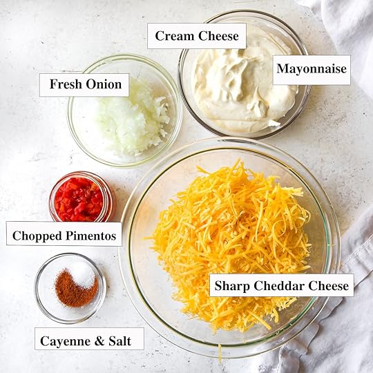 Overhead view of creamy pimento cheese spread thickly on toasted sourdough, with two glass jars of pimento cheese and scattered pretzels on a white surface.