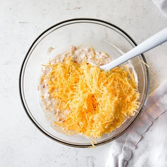Shredded sharp cheddar cheese being added to the pimento and cream cheese mixture in a glass bowl with a spatula, for homemade pimento cheese.