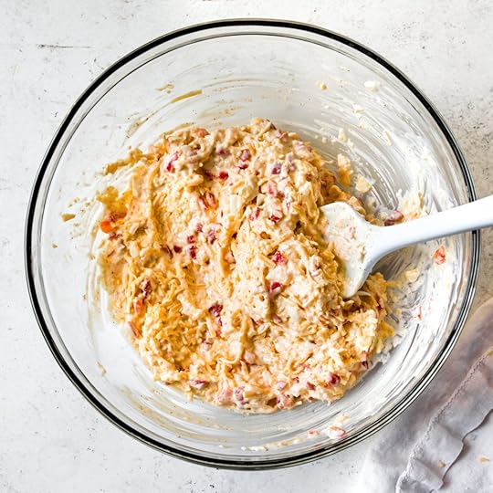 Fully mixed homemade pimento cheese in a glass bowl with a white spatula, showing shredded cheese and red pimentos.