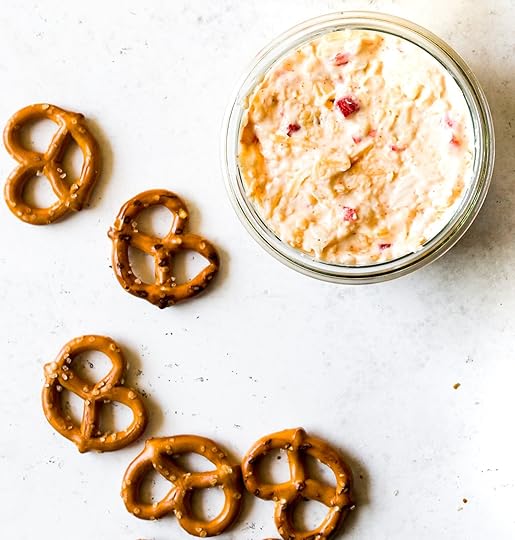 Creamy pimento cheese dip in a clear round jar on a light background, with scattered pretzel twists.