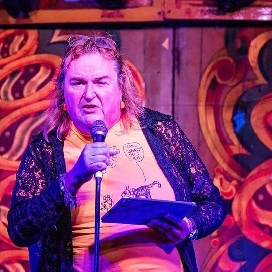 Sandra Bond reading her work aloud at an event, glasses perched on her head, witha microphone in one hand, an iPad in the other, and a determined look upon her face.