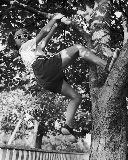 A black and white image of a girl smiling as she hangs from a tree branch, with one foot propped against the tree's trunk. She wears sunglasses