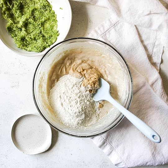 All-purpose flour being added to the wet batter for brown butter zucchini bread in a glass bowl with a spatula.