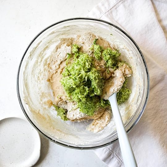 Shredded fresh zucchini added to brown butter zucchini bread batter in a glass mixing bowl with a white spatula.
