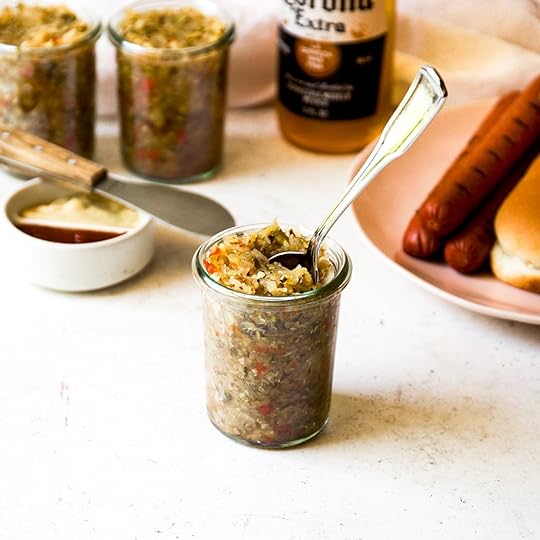 Jars of homemade spiced zucchini relish with a spoon, alongside hot dogs, buns, mustard, ketchup, and a bottle of beer in the background.