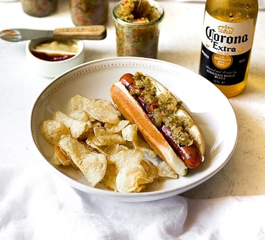 Close-up of a hot dog topped with homemade spiced zucchini relish and potato chips on a white plate, with jars of relish and a beer bottle in the background.