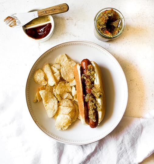Top-down view of a hot dog generously topped with homemade spiced zucchini relish, served with potato chips and a side of ketchup and mustard, with a jar of relish in the background.