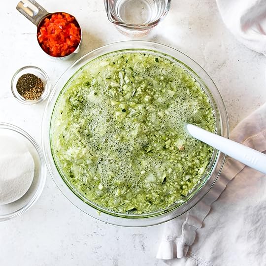 Large glass bowl with chopped zucchini and onions soaking in a brine solution, an early step in making homemade spiced zucchini relish, with red bell pepper and spices nearby.