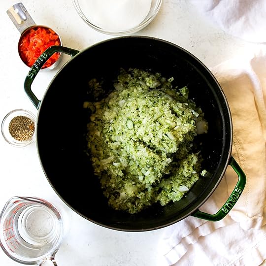 Drained zucchini and onion mixture placed in a dark green Dutch oven, ready for cooking the homemade spiced zucchini relish, with other ingredients like red pepper, sugar, and spices in the background.