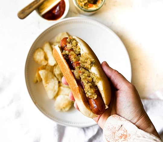A hand holding a hot dog topped with homemade spiced zucchini relish, with a plate of potato chips, condiments, and a jar of relish in the background.