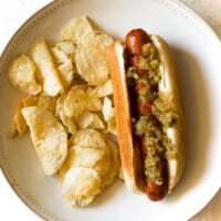Homemade spiced zucchini relish on a grilled hot dog with potato chips on a white plate.