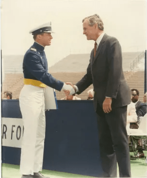 A young cadet in a military uniform shakes hands with a distinguished man in a suit during a graduation ceremony.