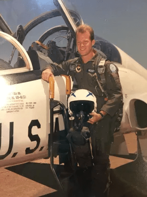 A smiling pilot in flight gear stands beside a U.S. Air Force training aircraft, holding a helmet and posing confidently.