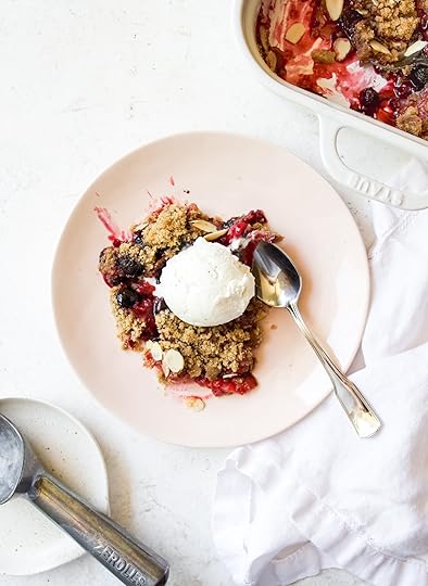 A serving of warm Mixed Berry Crumble with a scoop of vanilla ice cream on a pink plate, next to a white baking dish and an ice cream scoop.