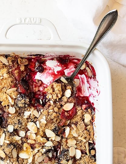 Close-up of a warm Mixed Berry Crumble in a white baking dish, showing the bubbling fruit filling, golden almond streusel topping, and a spoonful being served.