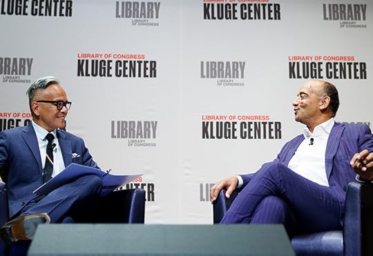 Medium two-shot of two men, both seated and wearing suits with white dress shirts, smiling and talking on stage.