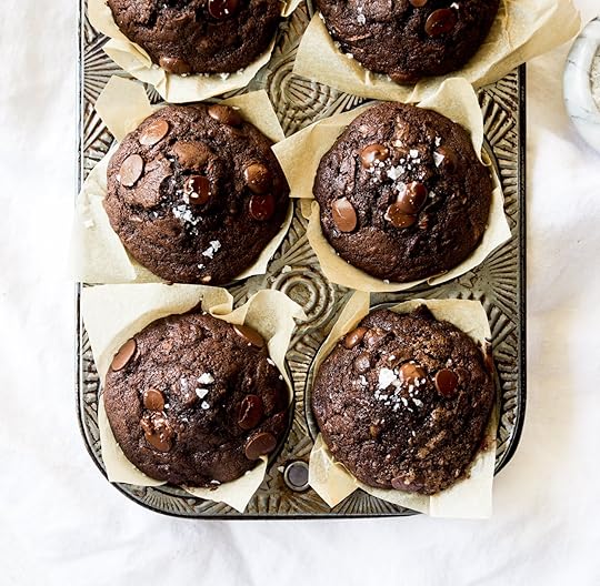 An overhead view of six bakery-style double chocolate zucchini muffins fresh from the oven in a rustic metal muffin tin. The rich, dark muffins are topped with melted chocolate chips and flaky sea salt and sit in crinkled parchment paper liners.