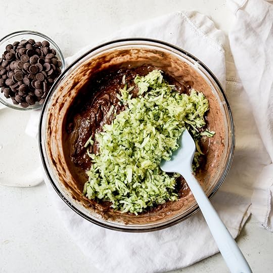 Overhead shot of a glass bowl with dark chocolate muffin batter, grated green zucchini, and a light blue spatula, with a small bowl of chocolate chips nearby on a white surface.