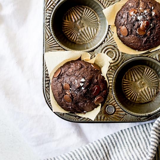 Overhead shot of two freshly baked dark double chocolate zucchini muffins with visible chocolate chips and flaky sea salt, cooling in a vintage metal muffin tin lined with parchment paper, on a white tablecloth with a striped towel.