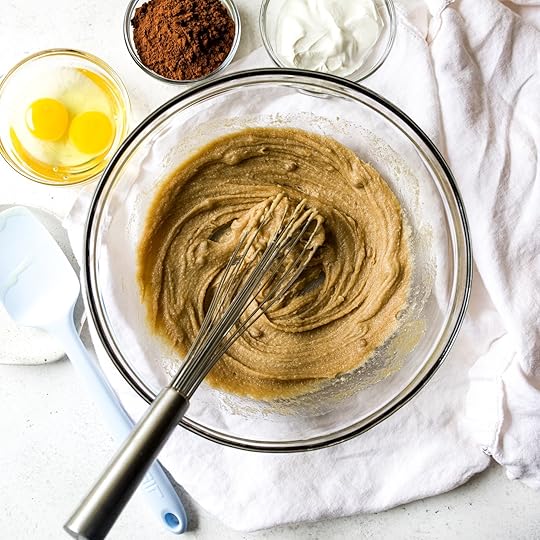 Overhead shot of light brown wet ingredients for double chocolate zucchini muffins being whisked in a clear glass bowl, with cocoa powder, sour cream, and eggs visible in the background.