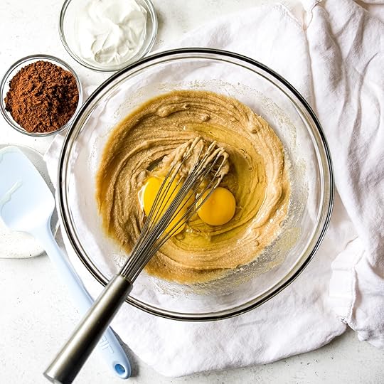 Overhead shot of two raw egg yolks and whites added to a creamy, whisked batter in a clear glass bowl, with a whisk resting inside and bowls of cocoa powder and sour cream in the background on a white surface.