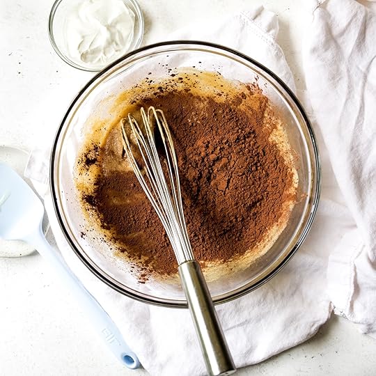 Overhead shot of rich unsweetened cocoa powder added to a light brown wet muffin batter in a clear glass bowl, with a whisk and a small bowl of sour cream visible on a white surface.