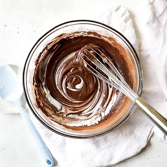 Overhead shot of rich dark chocolate muffin batter being swirled with creamy white sour cream in a clear glass bowl, with a metal whisk and blue spatula visible, illustrating the mixing process.