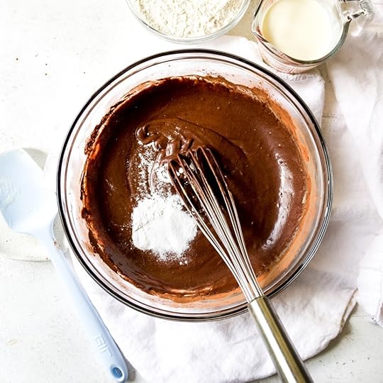 Overhead shot of white dry ingredients, likely baking powder, baking soda, and salt, being added to a rich, dark chocolate muffin batter in a clear glass bowl, with a whisk and other baking ingredients in the background.