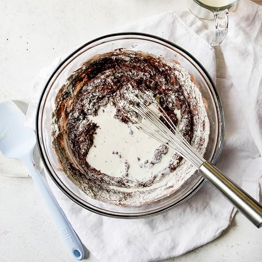An overhead shot of a glass bowl filled with chocolate zucchini muffin batter with flour on top and a whisk.
