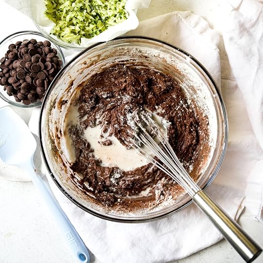 Overhead shot of rich dark chocolate muffin batter with unmixed white dry ingredients and a splash of milk being incorporated in a clear glass bowl with a whisk, showing the mixing process.