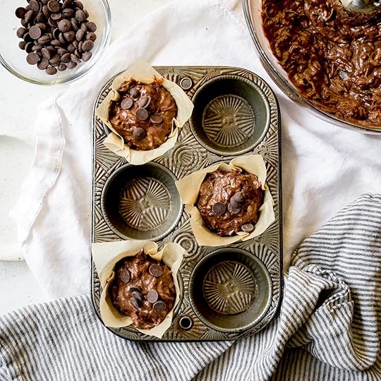 Overhead shot of rich double chocolate zucchini muffin batter with chocolate chips scooped into three parchment-lined cavities of a vintage metal muffin tin, ready for baking, with a bowl of remaining batter and chocolate chips nearby.