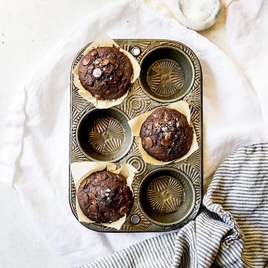 Overhead shot of three perfectly baked, rich double chocolate zucchini muffins with melted chocolate chips and a sprinkle of flaky sea salt, cooling in a vintage metal muffin tin on a white linen with a striped towel.