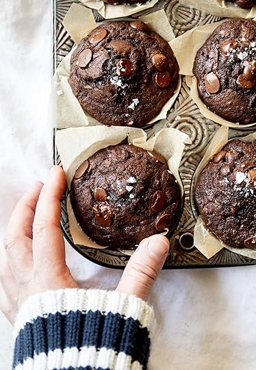 A hand in a striped sweater reaching for a warm, freshly baked double chocolate zucchini muffin with melted chocolate chips and a sprinkle of sea salt, nestled in a parchment liner in a rustic metal muffin tin.