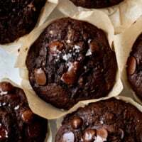 A close-up overhead shot of rich, bakery-style double chocolate zucchini muffins. The dark, moist muffins are topped with melted chocolate chips and a sprinkle of flaky sea salt and are sitting in rustic parchment paper liners.