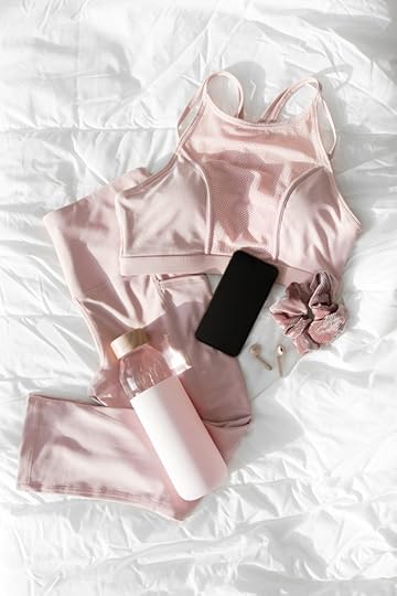Flat lay of pink workout gear, including a water bottle, wireless earbuds, a cell phone, and a hair scrunchy on a white duvet cover.